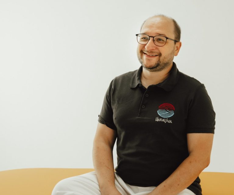 Über mich Markus Hayer von UndaViva sitzt freundlich auf einer Massageliege.